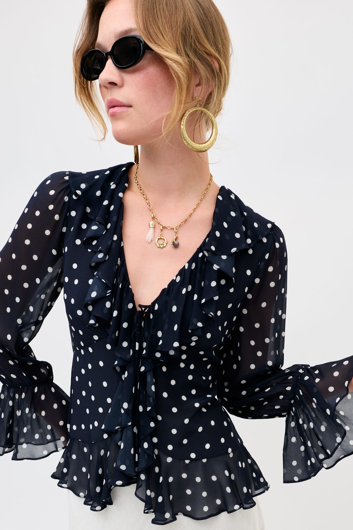 Woman wearing Sian - Gold hoop earrings and black oval sunglasses, styled with a navy blue sheer blouse with white polka dots and ruffled details, gazing to the side against a plain backdrop.