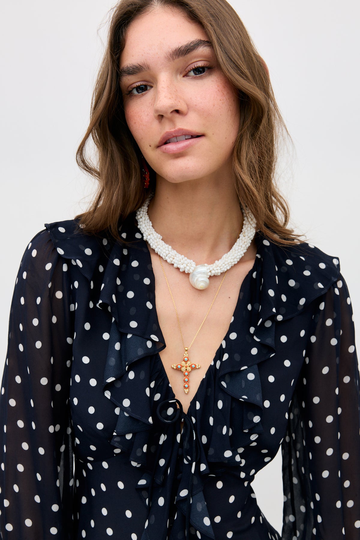 A woman with wavy brown hair wears the Ami - Layla Dot Navy blouse, styled with a layered pearl choker featuring a large bead and a gold cross pendant necklace, posing against a plain backdrop.