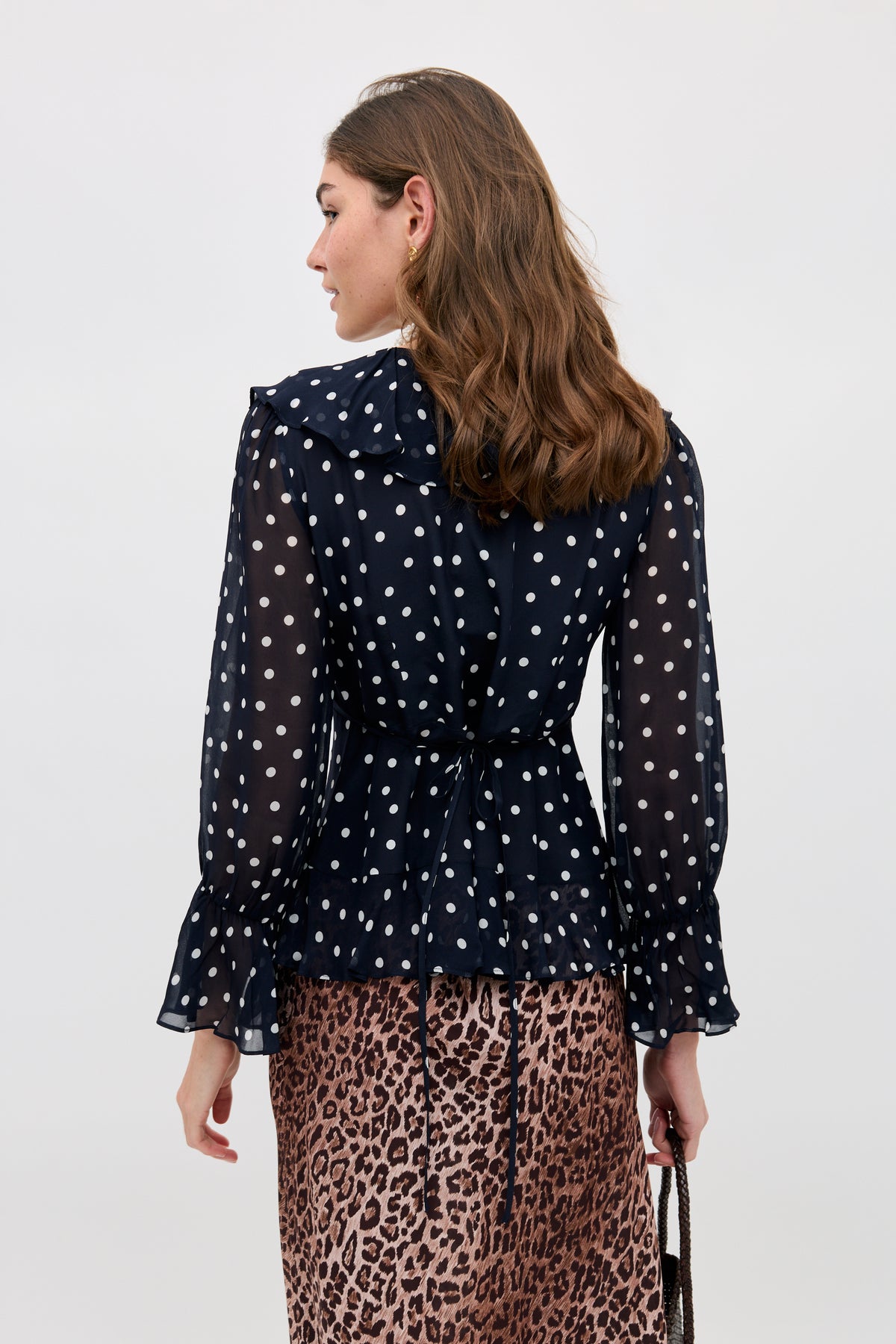 A woman with wavy brown hair is seen from behind wearing the Ami - Layla Dot Navy sheer blouse, featuring white polka dots and ruffled cuffs, paired with a leopard print skirt and holding a dark strap purse.