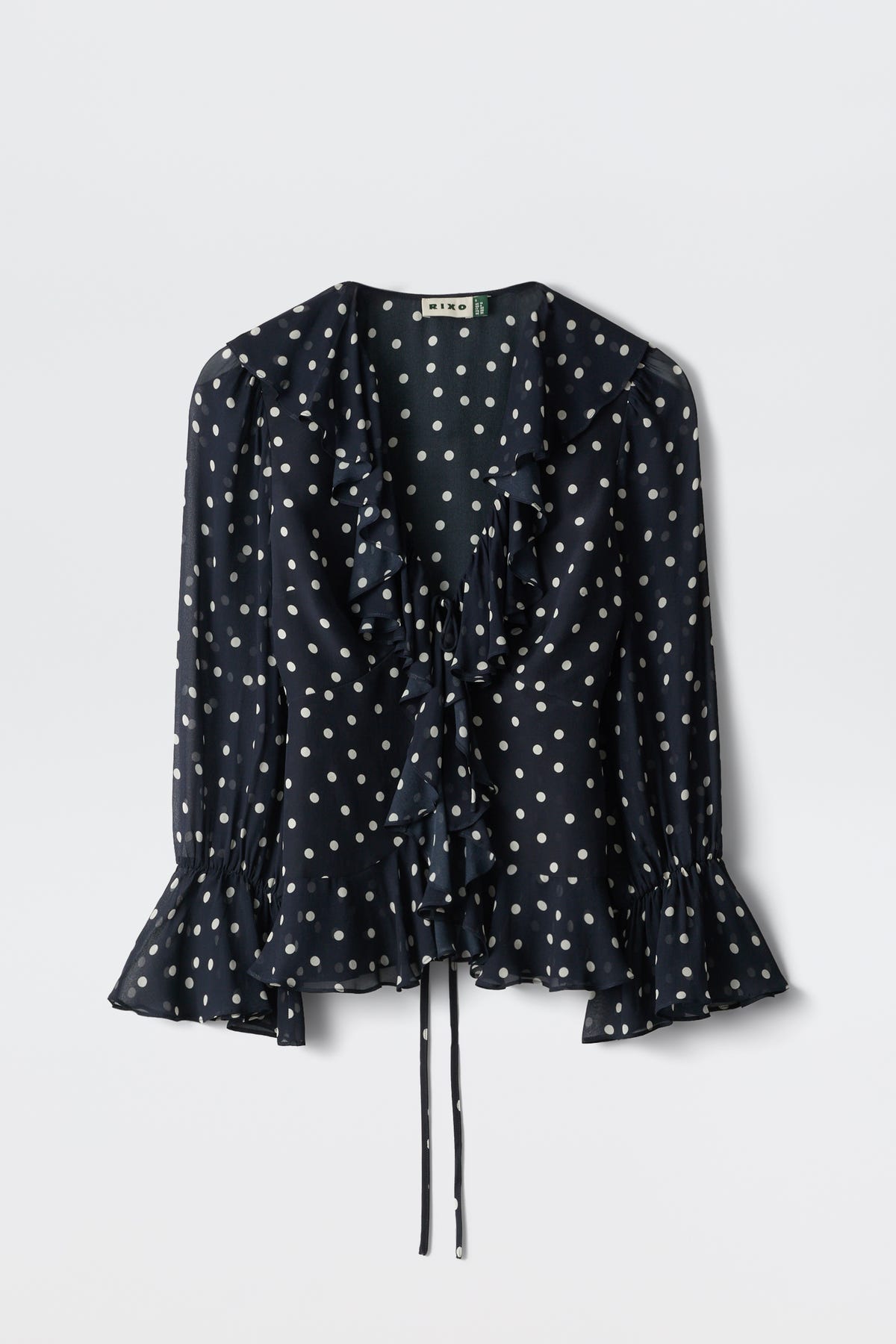 The Ami - Layla Dot Navy is a navy blue long-sleeve blouse featuring white polka dots, a ruffled neckline and cuffs, and a waist tie, shown against a plain white background.