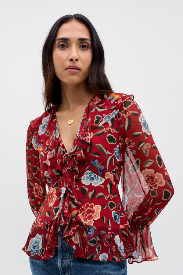 A woman models the Ami - Butterfly Floral Burgundy blouse, featuring sheer long sleeves, colorful floral print, waterfall ruffles, and a bow tie front. She pairs it with blue jeans and a gold necklace against a plain background.