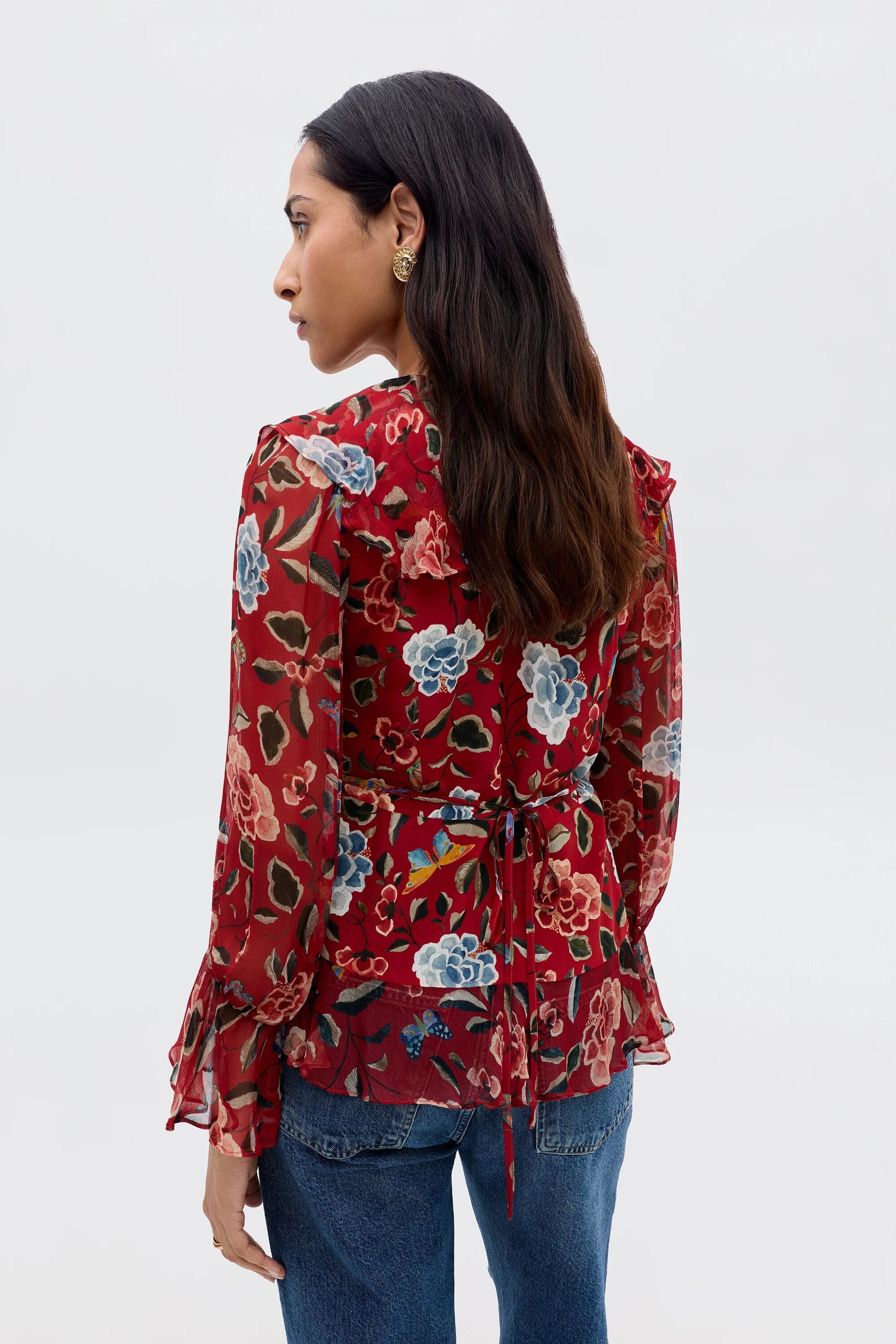 A woman with long dark hair, seen from behind, models the Ami - Butterfly Floral Burgundy blouse, featuring a colorful floral print, sheer fabric, waterfall ruffle cuffs, waist tie detail, and blue jeans.