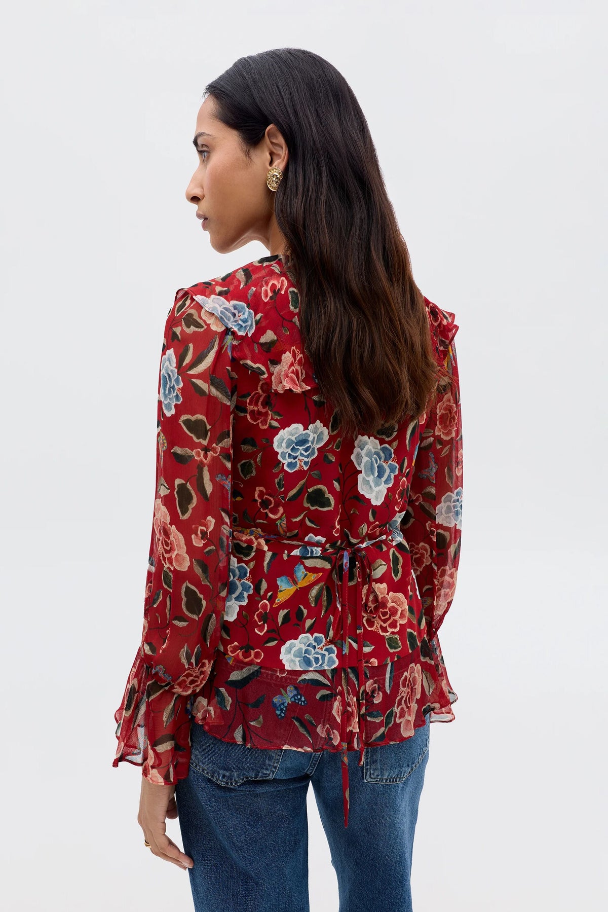 A woman with long dark hair, seen from behind, models the Ami - Butterfly Floral Burgundy blouse, featuring a colorful floral print, sheer fabric, waterfall ruffle cuffs, waist tie detail, and blue jeans.
