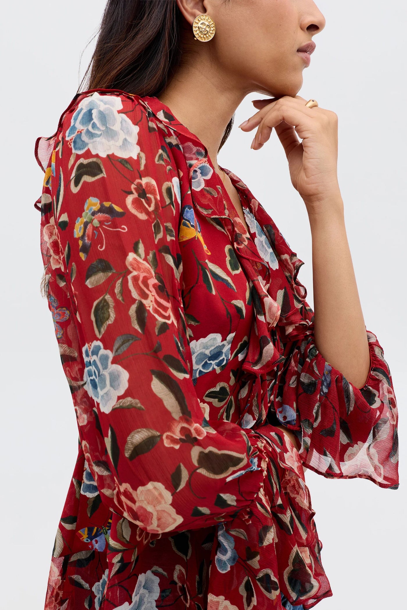 A woman models the Ami - Butterfly Floral Burgundy, an '80s-inspired long-sleeved blouse with waterfall ruffles, gold earrings, and a gold ring, posing with her hand on her chin against a plain light background.