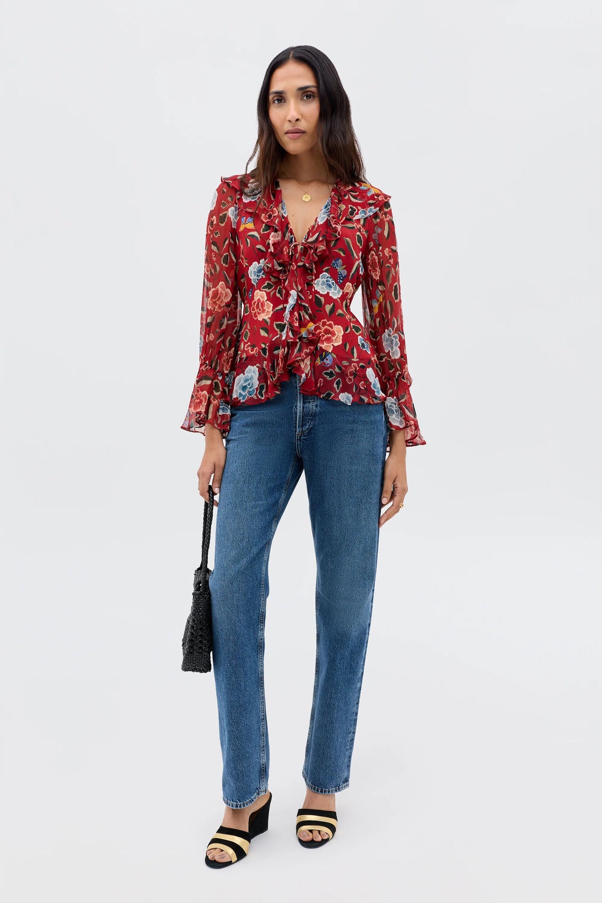 A woman with long dark hair and a neutral expression stands against a plain background wearing the Ami - Butterfly Floral Burgundy blouse, blue jeans, black sandals, and holding a black woven bag.