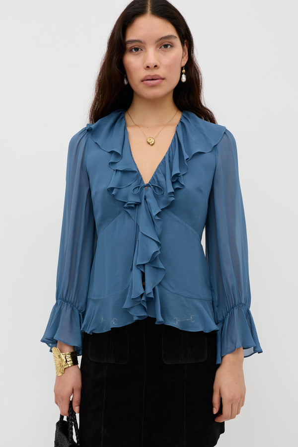 A woman with long dark hair wears the Ami - Stone Blue blouse with ruffled details and sheer sleeves, paired with a black skirt, gold jewelry, and a black bag. She stands against a plain white background, looking at the camera.
