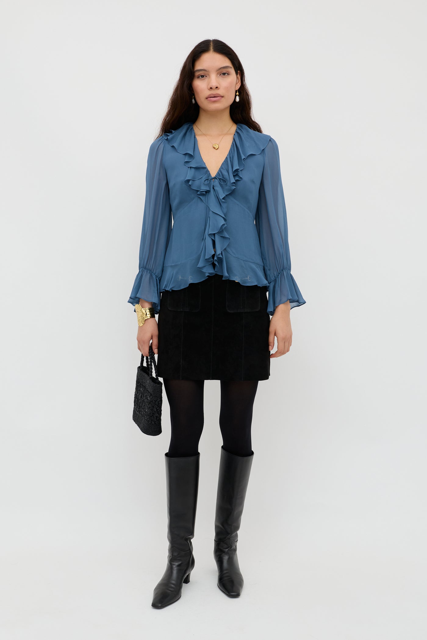 A woman stands against a white background wearing the Ami - Stone Blue ruffled blouse, a black mini skirt, black tights, knee-high boots, gold jewelry, and holding a small black textured handbag.