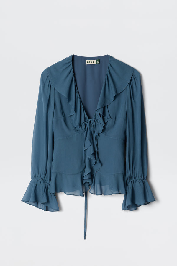 The Ami – Stone Blue is a long-sleeved blouse featuring a ruffled collar, ruffled front, tie neckline, and flared cuffs in deep blue, shown against a plain white background.