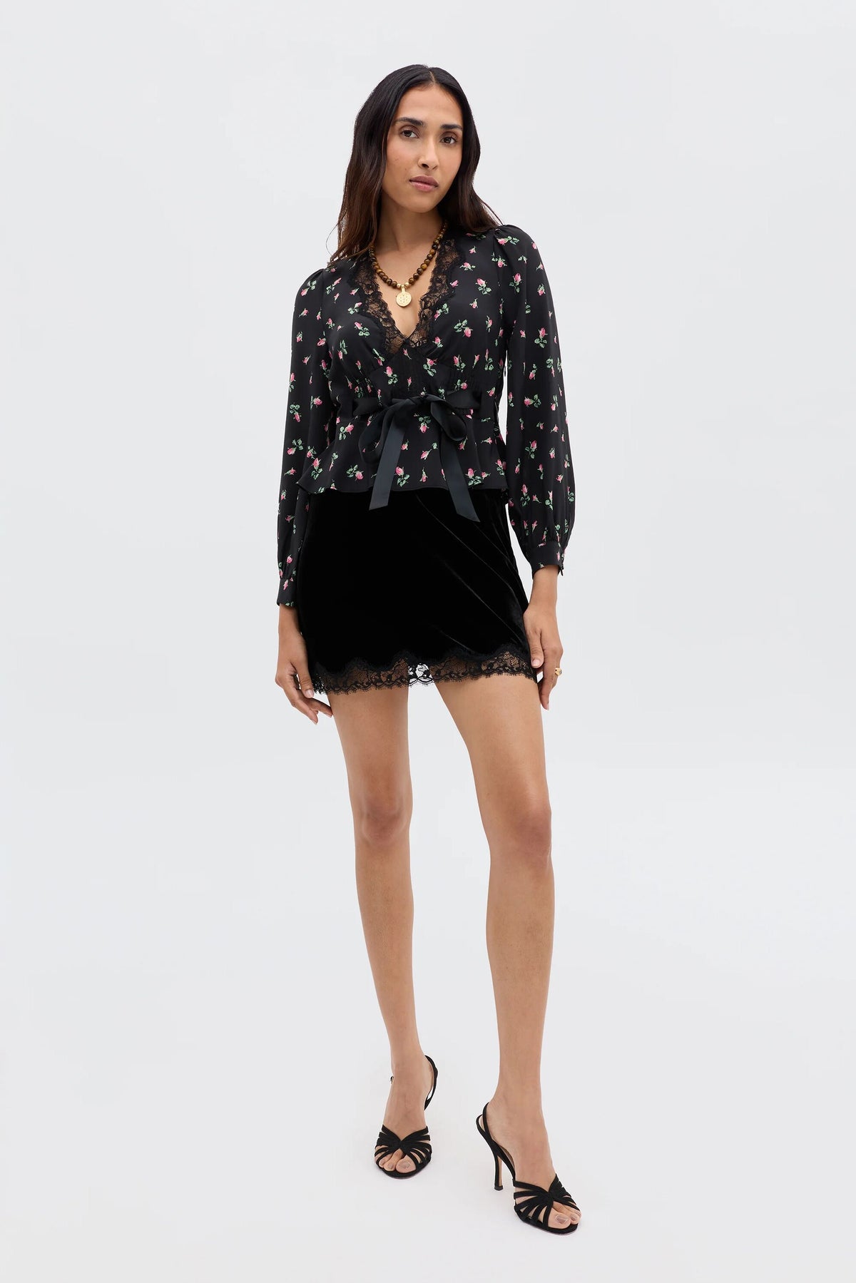 A woman exudes romantic femininity in the RIXO Charis - Sienna Bud Black blouse with a belted waist, paired with a black velvet mini skirt featuring lace trim. She styles her long hair with black heels and layered necklaces.