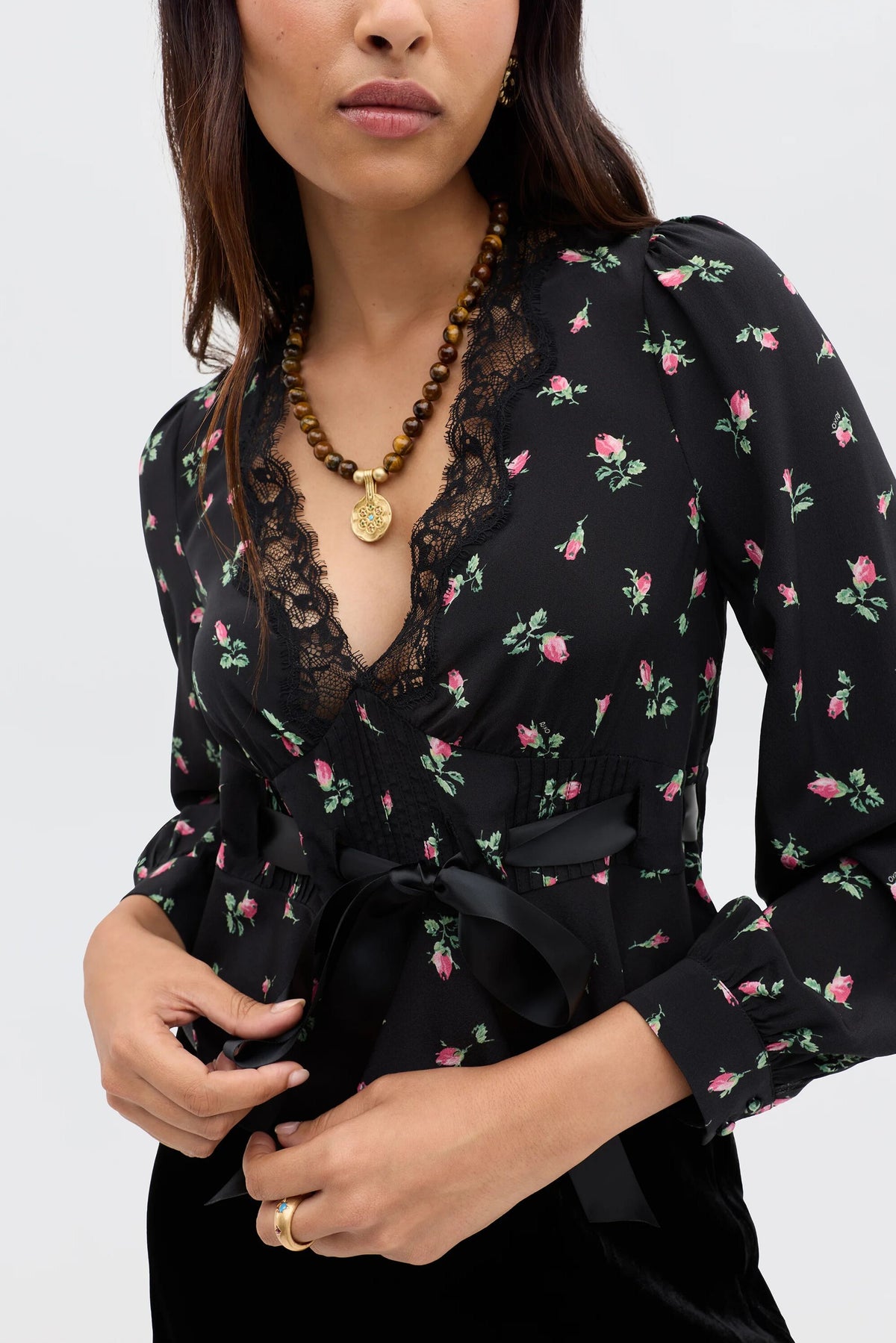 Embodying romantic femininity, a woman wears the Charis - Sienna Bud Black blouse with a lace-trimmed neckline and satin ribbon belt, paired with a beaded gold pendant necklace and gold hoop earrings—a signature RIXO-inspired look.