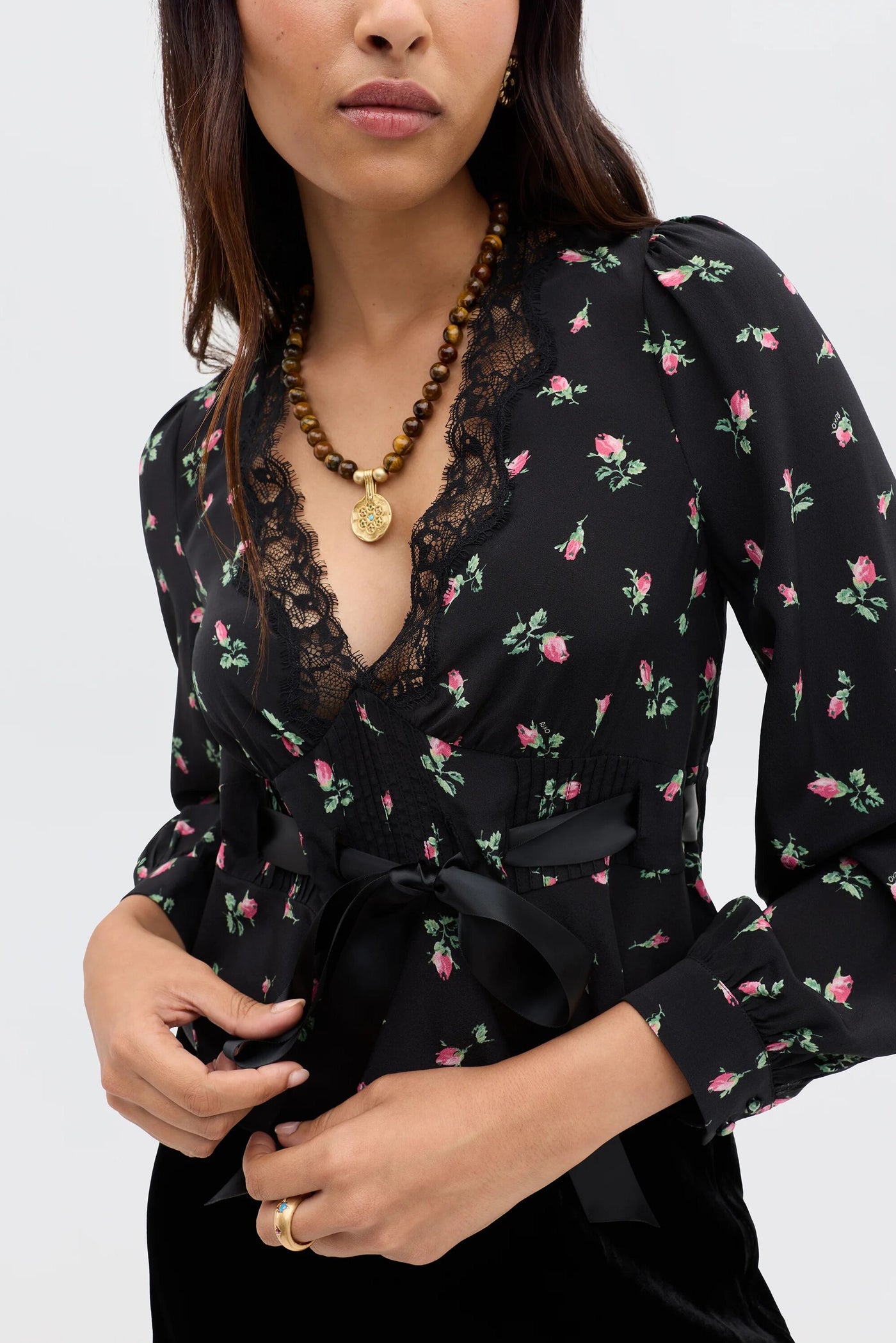 Embodying romantic femininity, a woman wears the Charis - Sienna Bud Black blouse with a lace-trimmed neckline and satin ribbon belt, paired with a beaded gold pendant necklace and gold hoop earrings—a signature RIXO-inspired look.