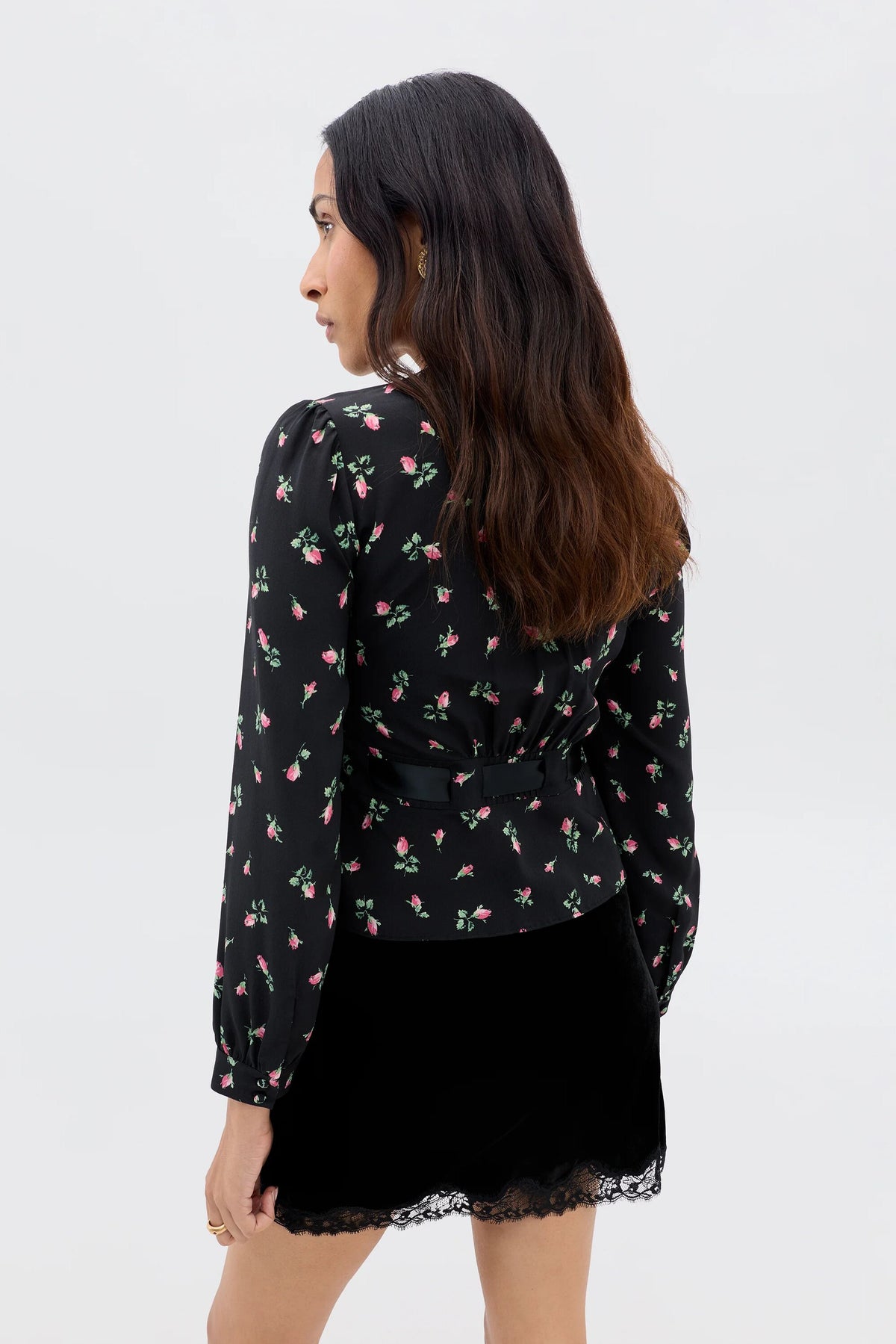Exuding romantic femininity, a woman with long wavy hair faces away, wearing the Charis - Sienna Bud Black blouse with puffed sleeves and a black skirt with lace trim against a plain light background.