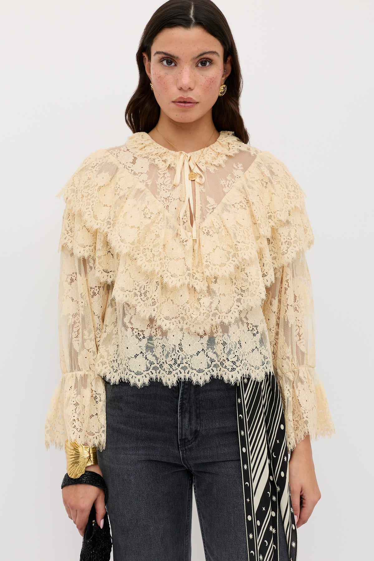 A woman wearing the Faryn - Vintage Cream sheer lace blouse with ruffles, paired with black jeans and a black-and-white patterned scarf hanging from her waist, stands against a plain white background.