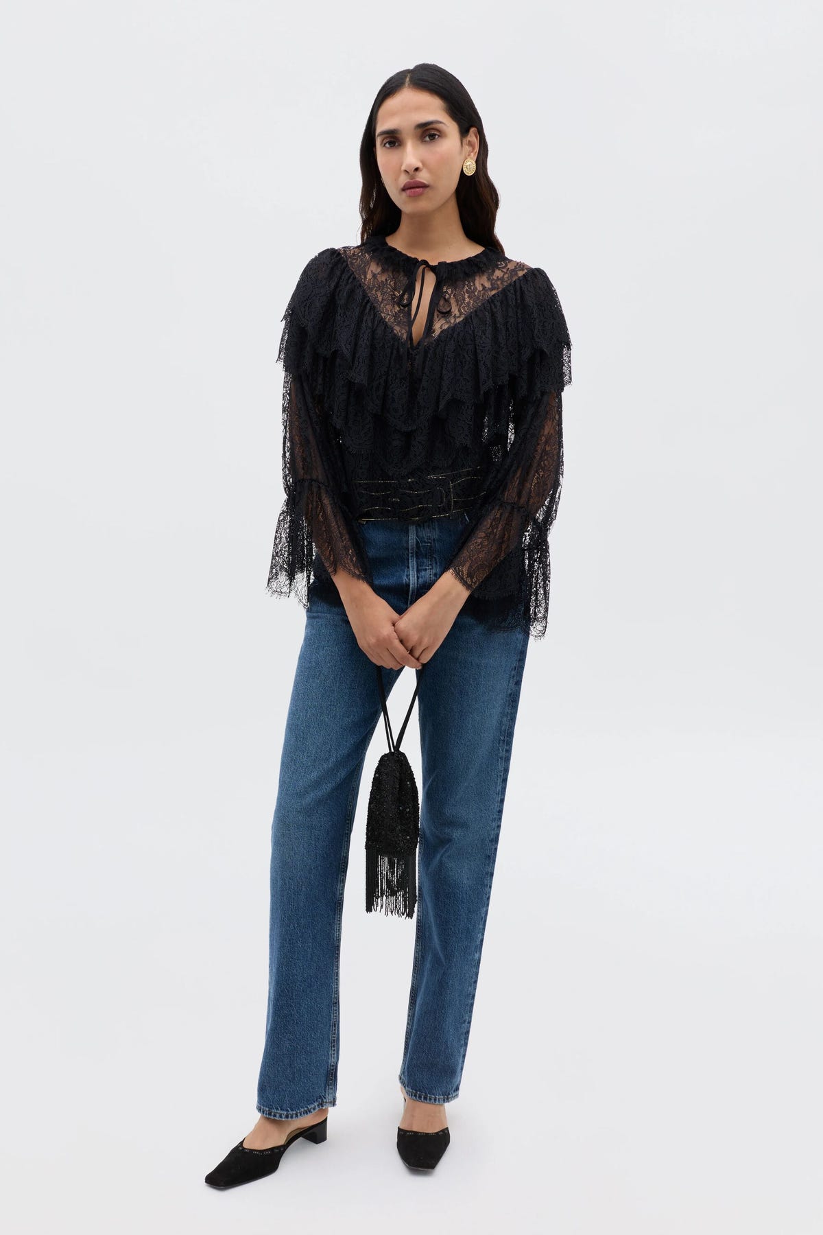 A woman stands against a plain background wearing the Faryn - Black lace ruffle blouse with blue jeans, black shoes, and holding a small black fringed handbag.