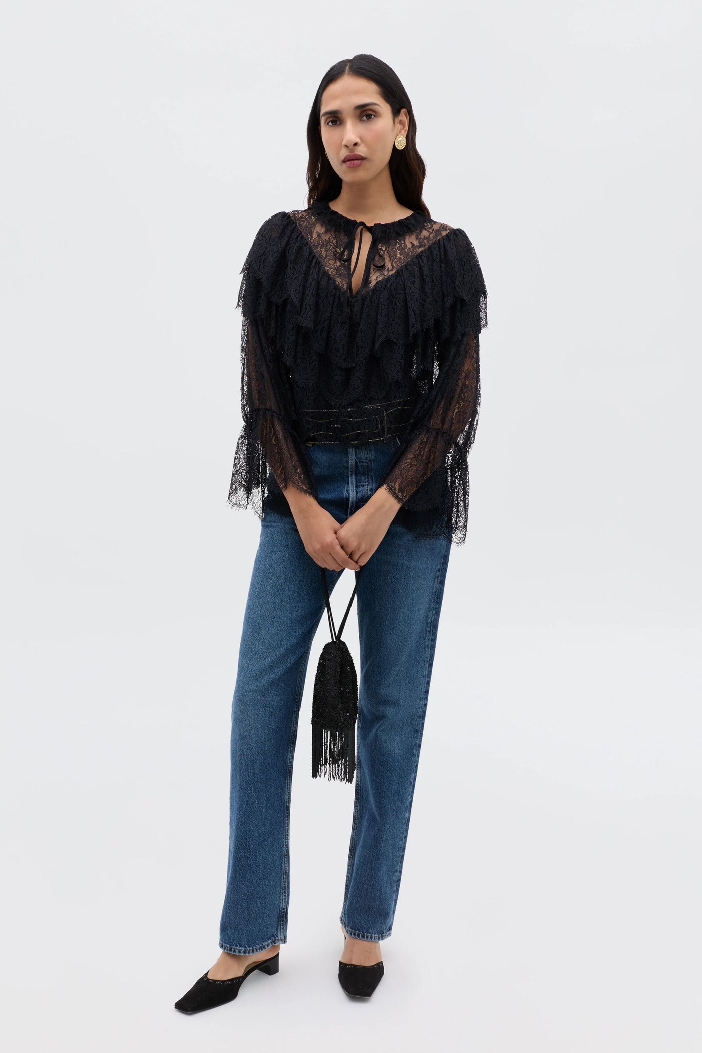A woman stands against a plain background wearing the Faryn - Black lace ruffle blouse with blue jeans, black shoes, and holding a small black fringed handbag.