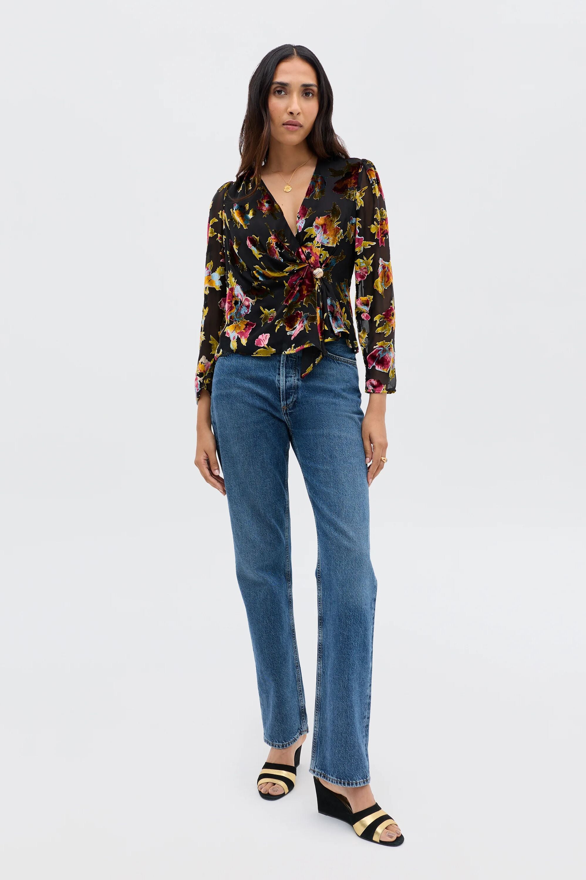 A woman with long, dark hair and a neutral expression stands against a plain background wearing the Everlyn - Countryside Devoré Black blouse, paired with blue straight-leg jeans and black open-toe sandals.
