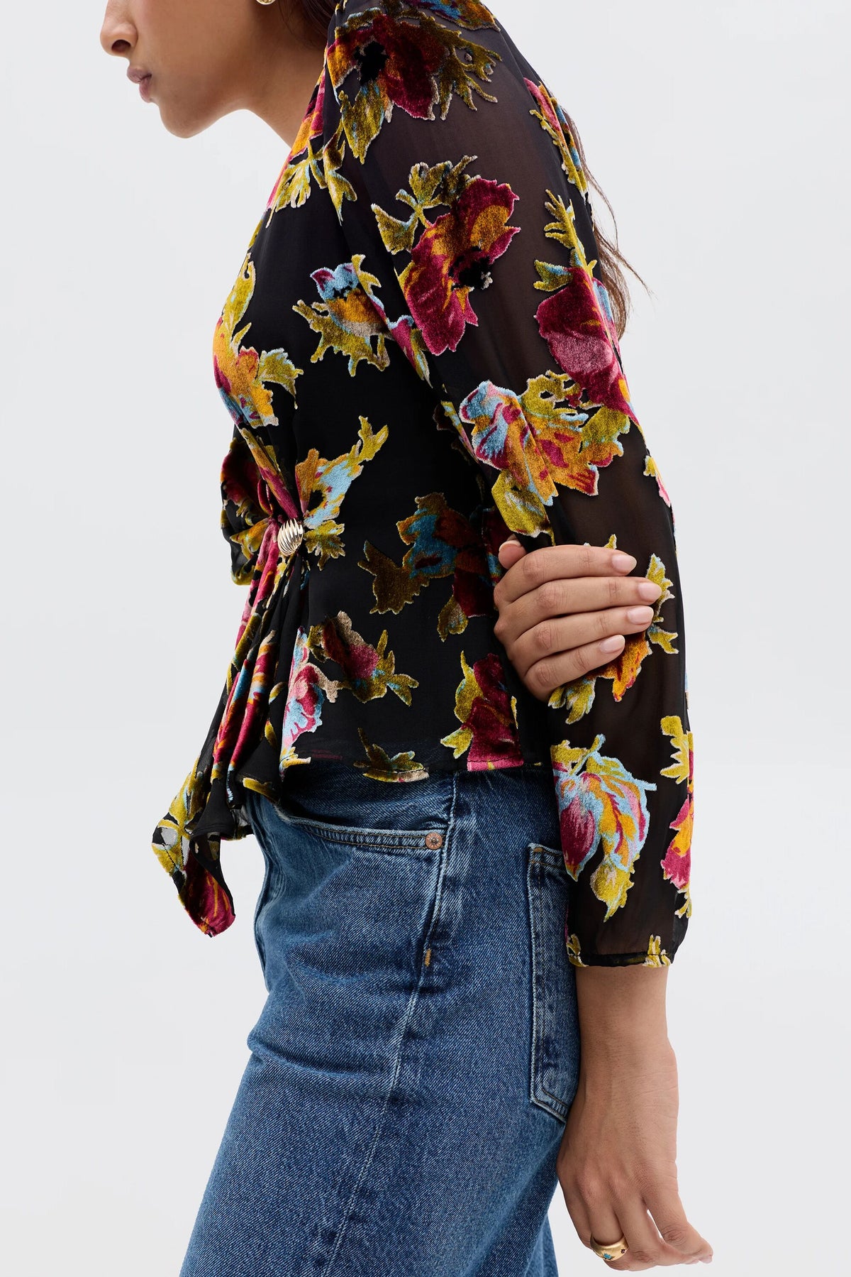 A woman, shown from shoulders down in side profile with crossed arms, wears the Everlyn - Countryside Devoré Black: a sheer black blouse with colorful floral prints and long sleeves, styled with blue denim jeans.