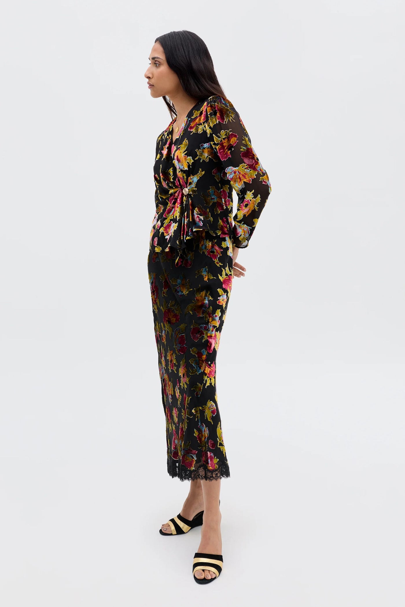 A woman with long dark hair stands sideways against a plain light background, wearing the Everlyn - Countryside Devoré Black dress with vintage-inspired floral top, lace hem, and long sleeves, paired with black and beige sandals.