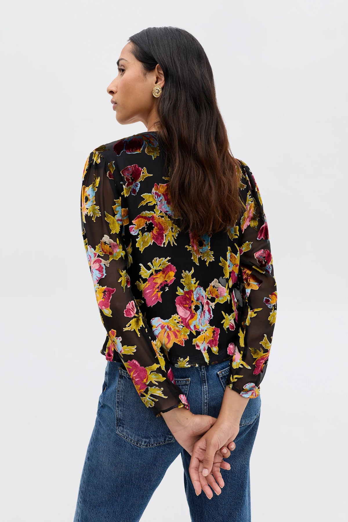 A woman stands facing away, head turned to the side, wearing the Everlyn - Countryside Devoré Black floral top with sheer black fabric and blue jeans. Her hands are clasped behind her back against a plain light background.