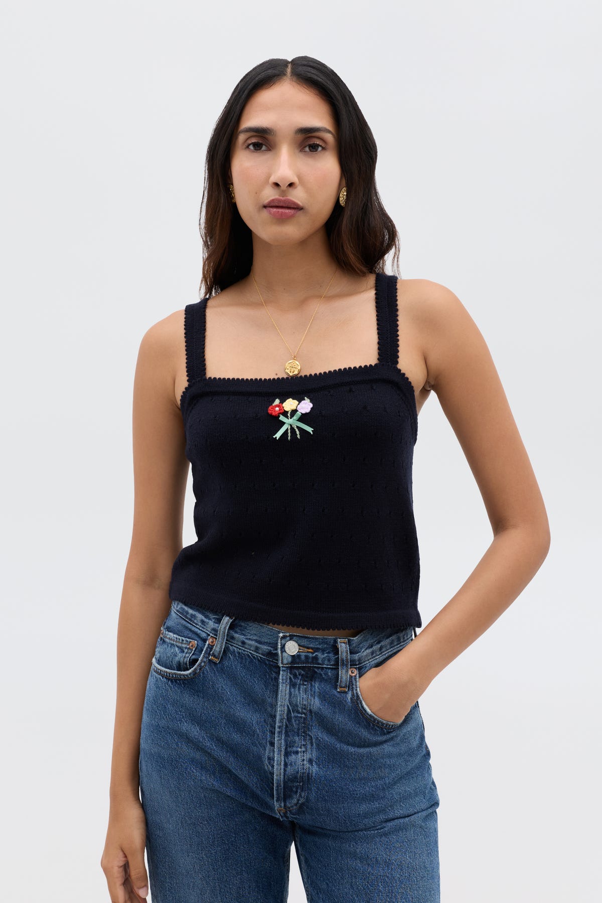 A woman with long brown hair wears the Perla - Floret Embroidery Deep Navy sleeveless, square-neck top paired with a cropped merino wool sweater, gold necklace, and blue jeans, standing against a plain light background.