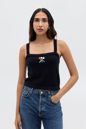 A woman with long brown hair wears the Perla - Floret Embroidery Deep Navy sleeveless, square-neck top paired with a cropped merino wool sweater, gold necklace, and blue jeans, standing against a plain light background.