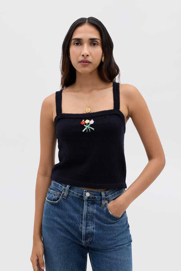 A woman with long brown hair wears the Perla - Floret Embroidery Deep Navy sleeveless, square-neck top paired with a cropped merino wool sweater, gold necklace, and blue jeans, standing against a plain light background.