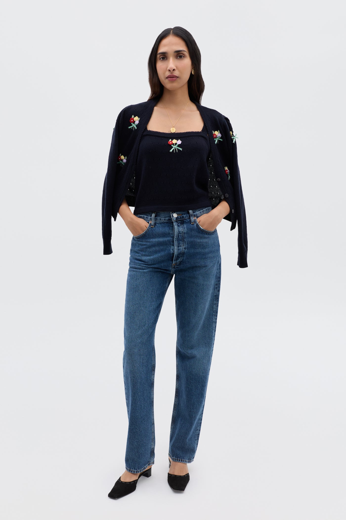 A woman with long dark hair stands against a plain background, wearing the Perla - Floret Embroidery Deep Navy twinset, blue jeans, and black shoes, with her hands in her jean pockets.