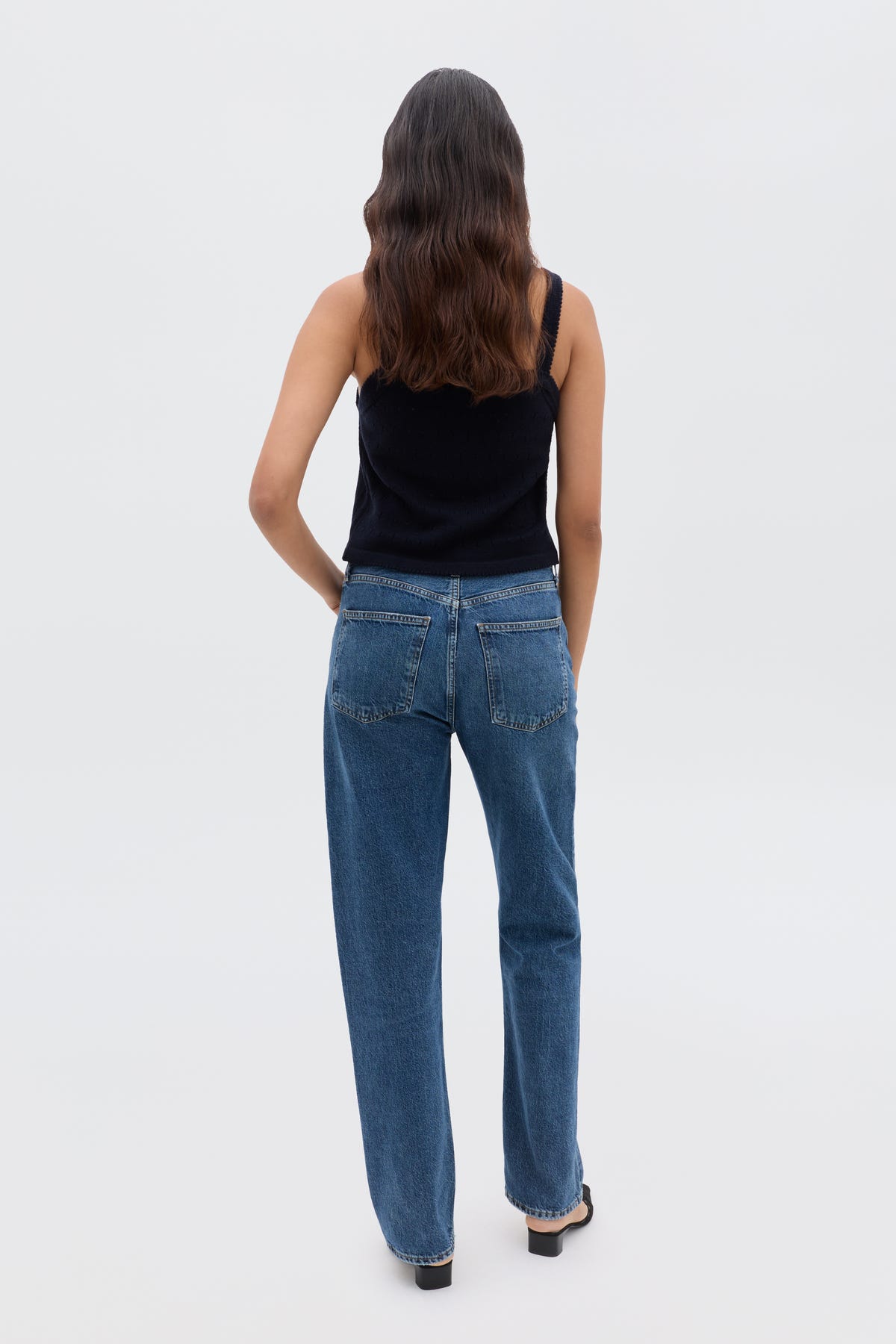 A person with long, wavy brown hair stands facing away, wearing the Perla - Floret Embroidery Deep Navy sweater with blue jeans and black shoes against a plain light background.
