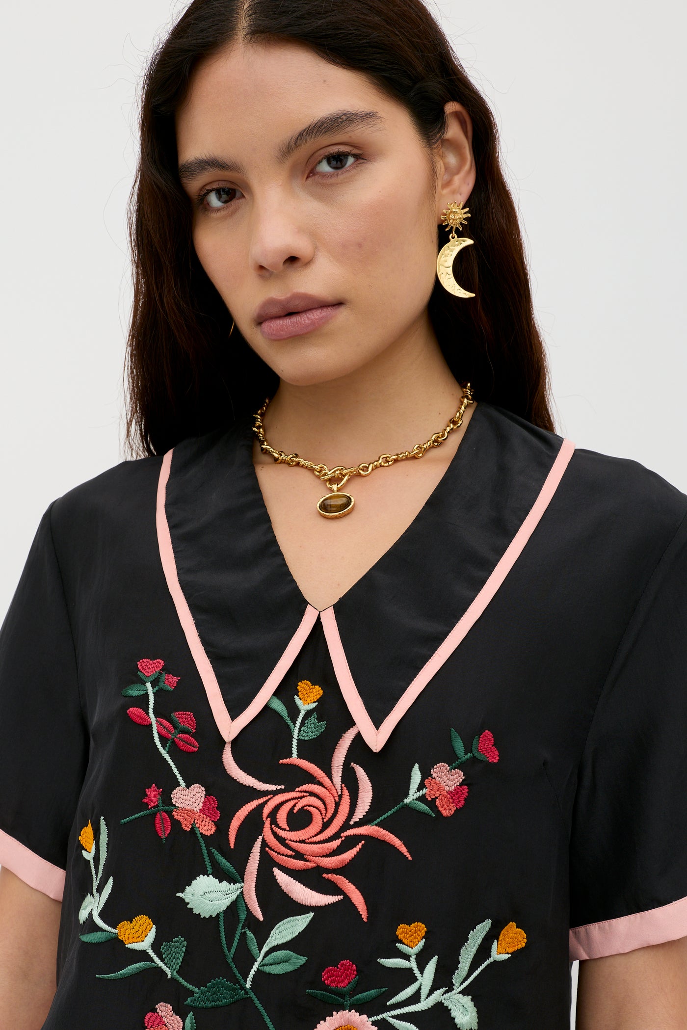 A woman with long dark hair wears the Nicolina - Frida Embroidery Black blouse with pink trim and colorful floral details, gold statement earrings (one crescent moon-shaped), and a gold chain necklace, gazing neutrally into the camera.