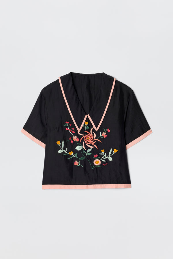 The Nicolina - Frida Embroidery Black is a vintage-inspired blouse with short sleeves, a deep V-neck trimmed in light pink, pink sleeve edging, and vibrant floral embroidery on the front.