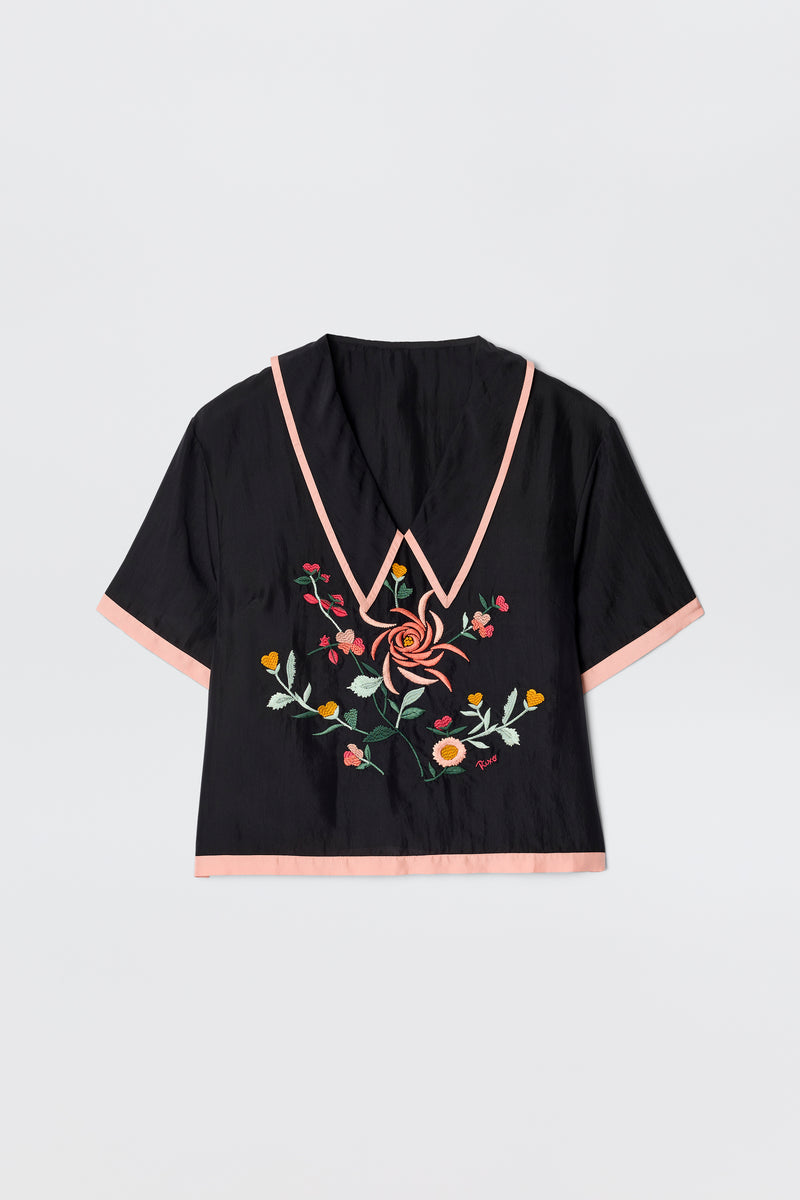 The Nicolina - Frida Embroidery Black is a vintage-inspired blouse with short sleeves, a deep V-neck trimmed in light pink, pink sleeve edging, and vibrant floral embroidery on the front.