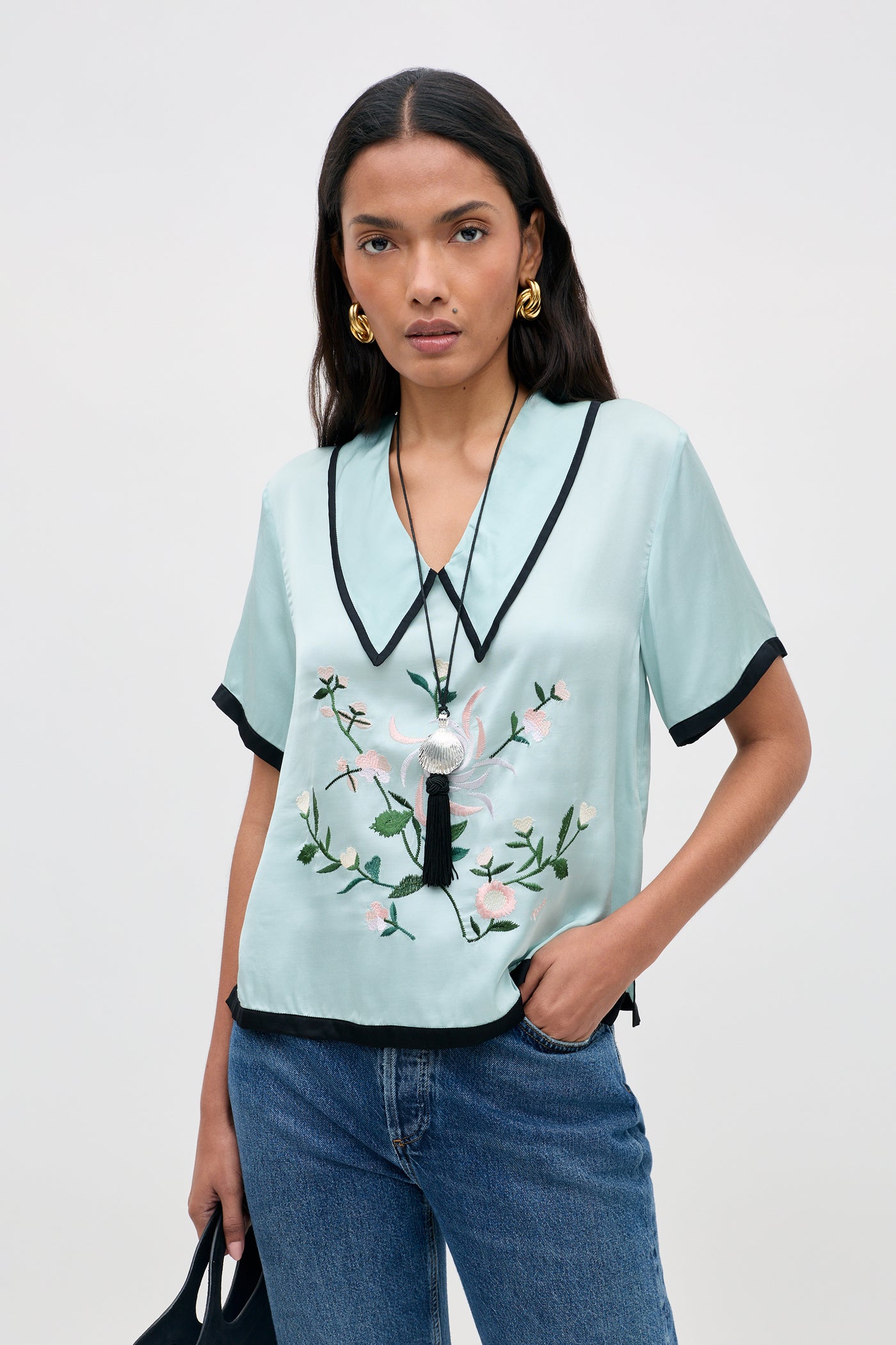A woman with long dark hair models the Nicolina - Frida Embroidery Mist light blue satin blouse featuring black trim and floral embroidery, styled with blue jeans. She accessorizes with gold earrings and a pendant necklace, standing against a plain light background.