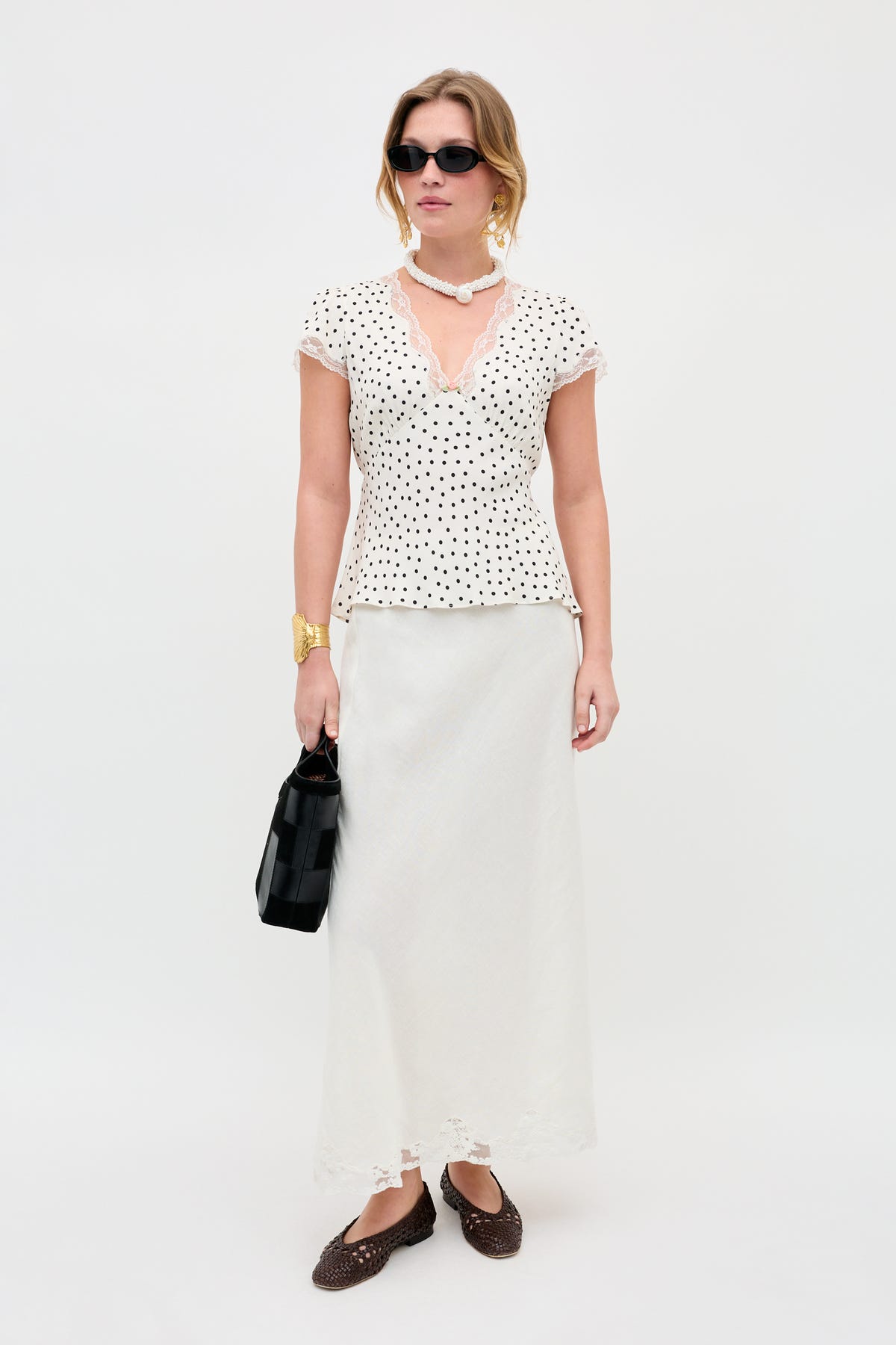 A woman stands against a white background wearing the Karia - Layla Dot Ivory blouse, paired with a long white skirt, brown woven shoes, black sunglasses, gold jewelry, a black bag, and a white beaded necklace.