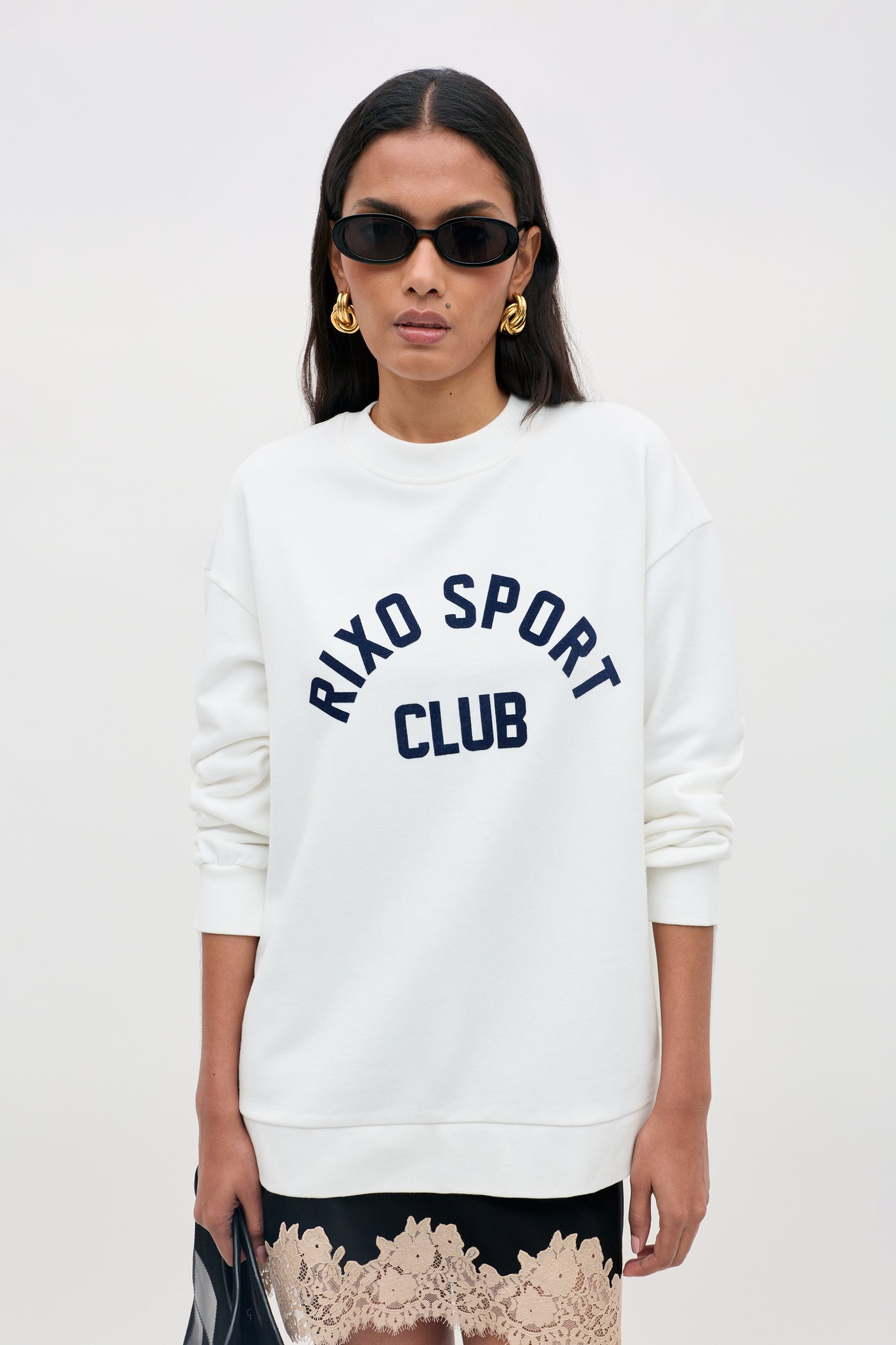 A woman wearing sunglasses and gold earrings models the Blaze - Rixo Sport Club White sweatshirt, paired with a lace-trimmed skirt and holding a bag.