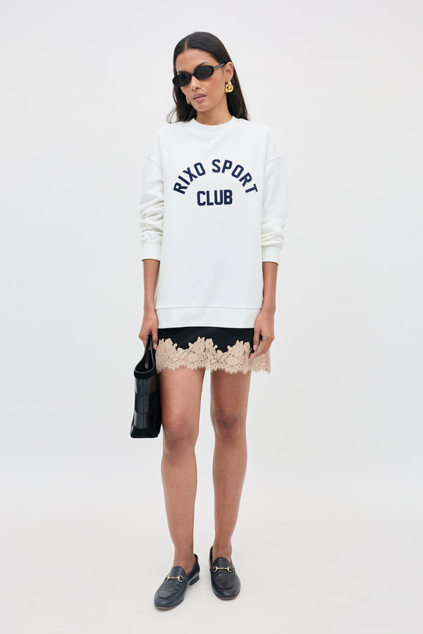 Wearing the Blaze - Rixo Sport Club White sweatshirt, a woman stands against a plain background styled with sunglasses, a black skirt featuring a beige lace hem, black loafers, gold earrings, and carrying a black handbag.