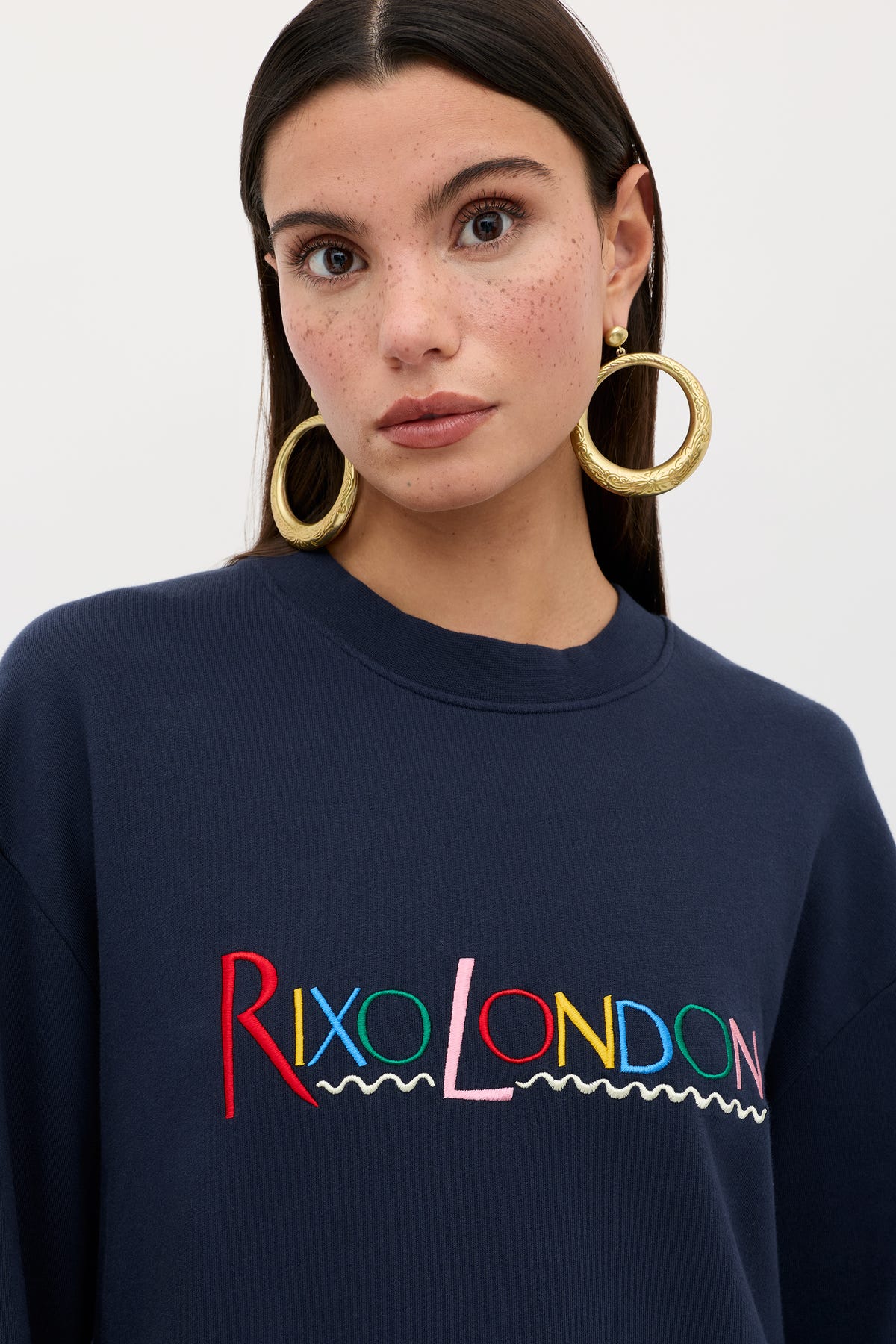 A woman with long dark hair, freckled skin, and gold hoop earrings wears the Blaze - London Embroidery Navy sweatshirt. "London" is embroidered in colorful letters. She stands against a simple light background.