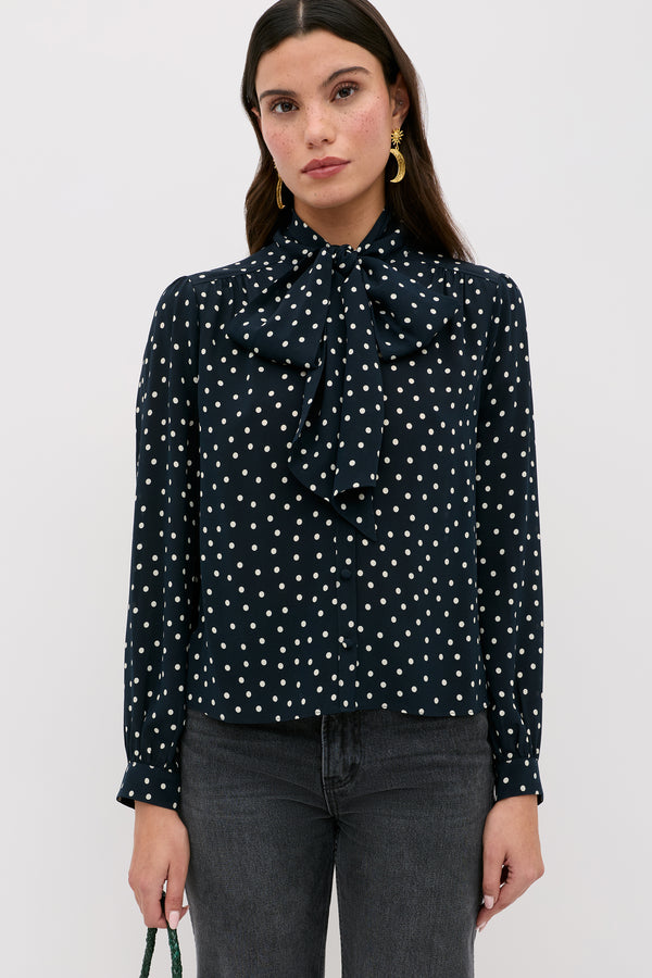 A woman with long dark hair and a neutral expression stands facing forward, wearing the Melaina - Layla Dot Navy top with white polka dots, paired with dark jeans, gold earrings, and carrying a green bag.
