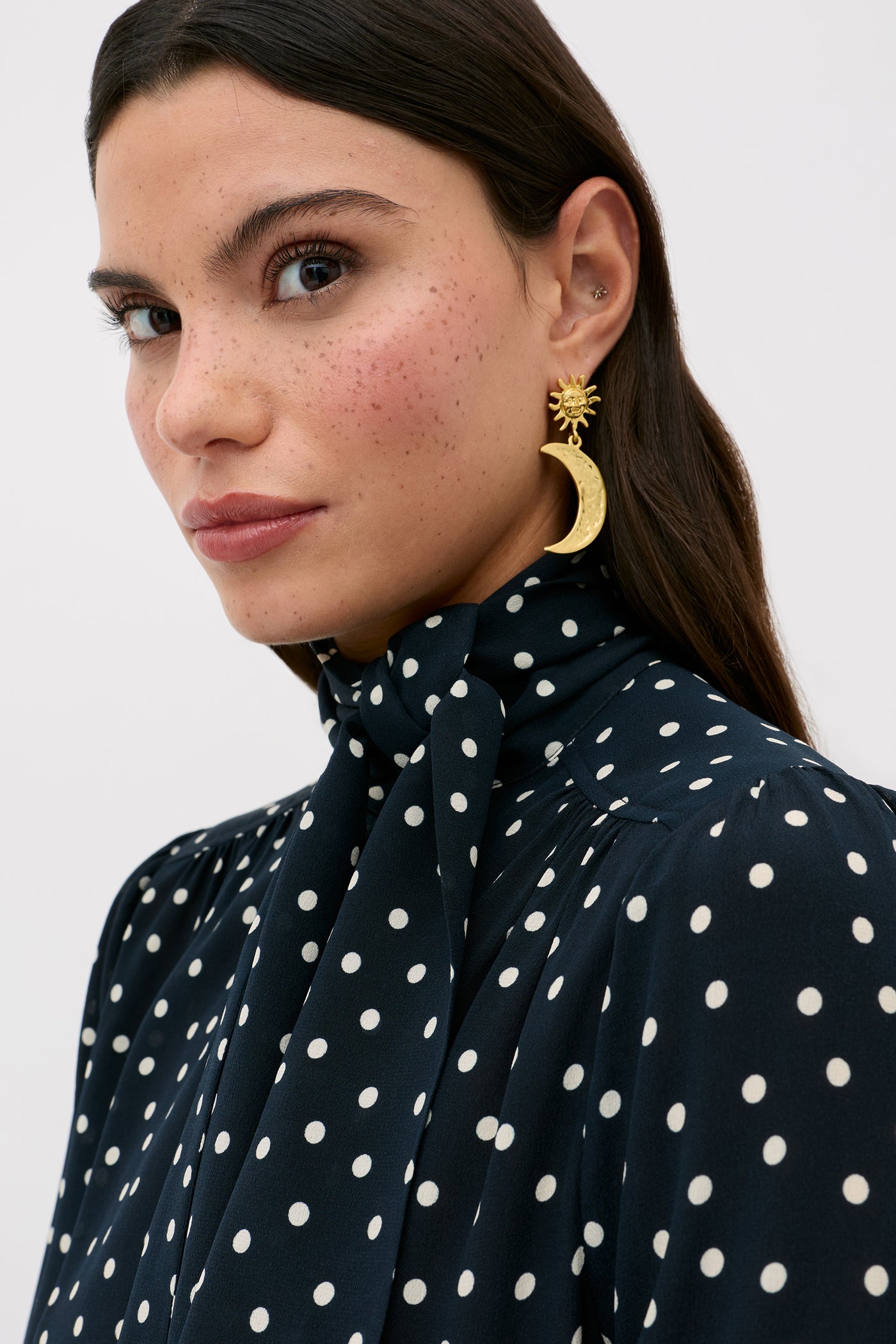 A woman with long brown hair wears the Melaina - Layla Dot Navy, a navy blouse with white polka dots and a scarf neckline. She has freckles, subtle makeup, and gold crescent moon and sun earrings, gazing to the side.