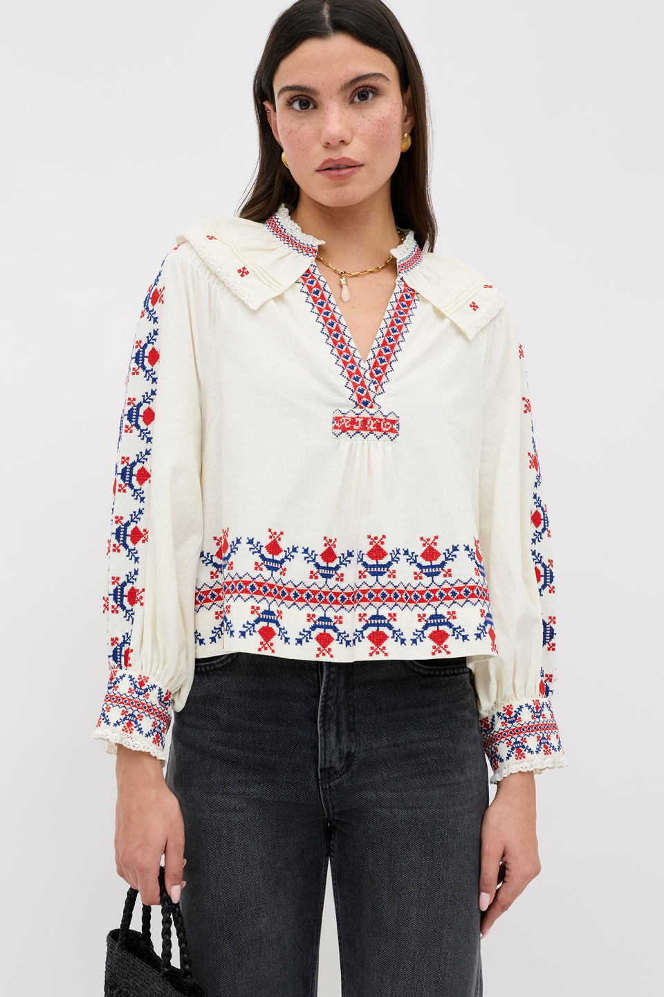 A woman wears the Brisella Gea Geo Embroidery Cream blouse—cream with red and blue floral embroidery, long sleeves, V-neck with ruffle trim—paired with black jeans, standing against a plain white background.