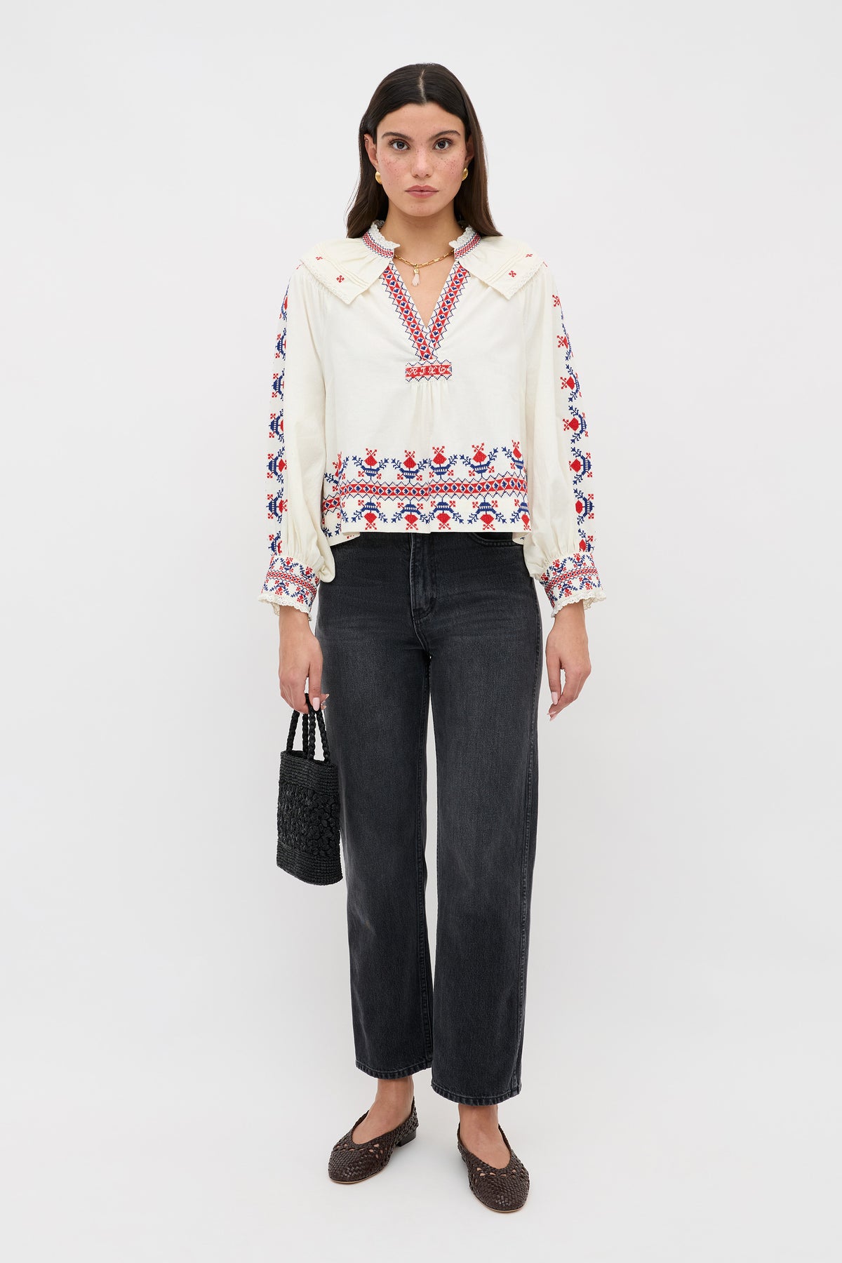 A woman stands against a white background wearing the Brisella - Gea Geo Embroidery Cream blouse, black wide-leg jeans, brown woven shoes, and holding a small black textured handbag.