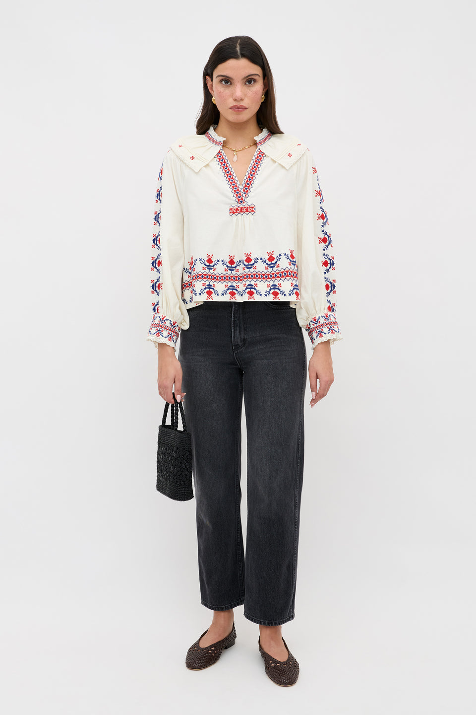 A woman stands against a white background wearing the Brisella - Gea Geo Embroidery Cream blouse, black wide-leg jeans, brown woven shoes, and holding a small black textured handbag.