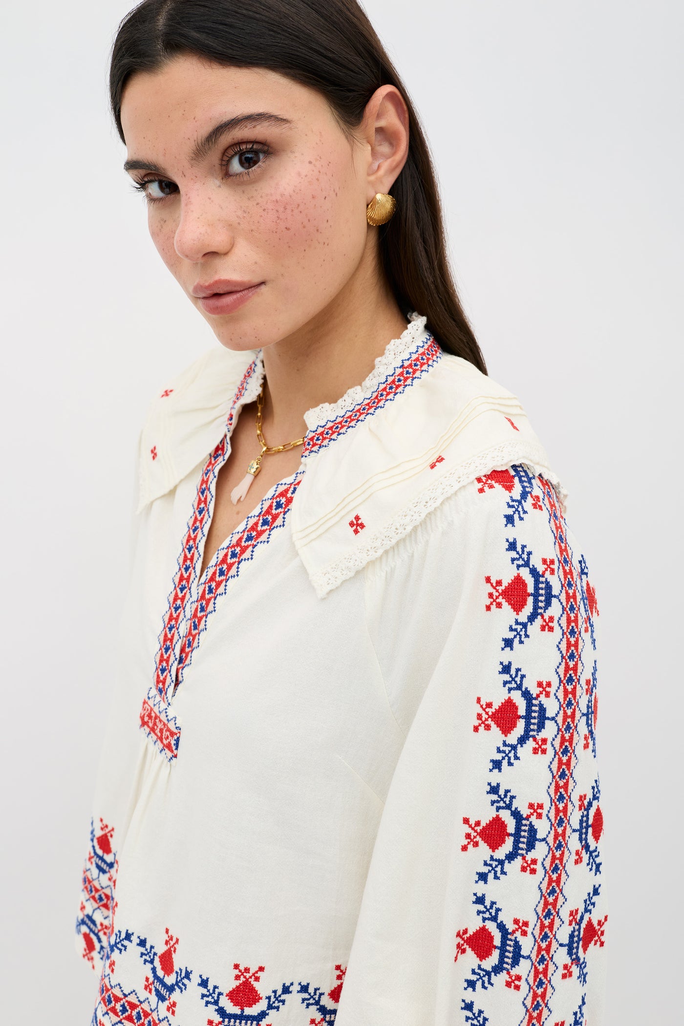 A woman with dark hair, freckles, and gold earrings models the Brisella - Gea Geo Embroidery Cream blouse, featuring blue and red embroidered patterns, against a plain light background.