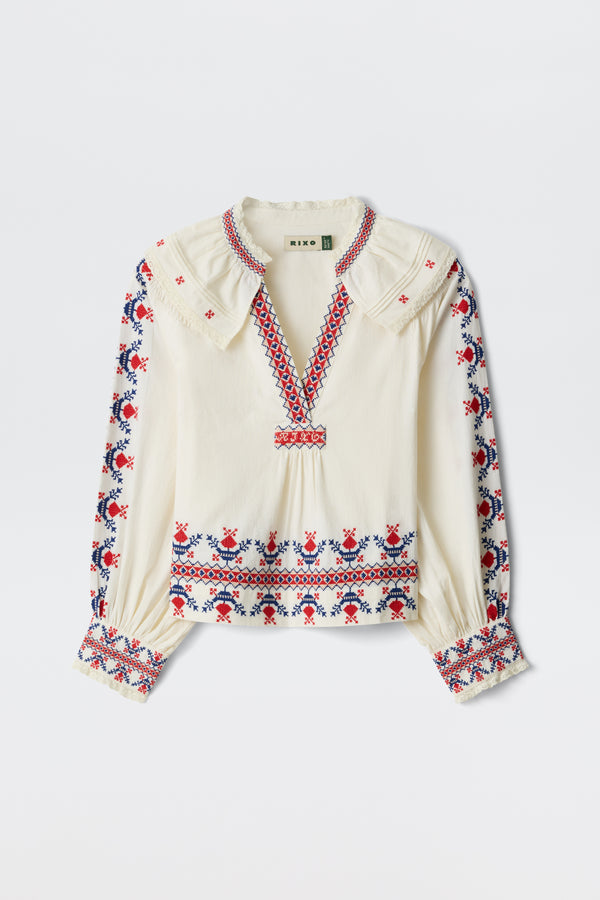 The Brisella - Gea Geo Embroidery Cream blouse features a cream color, long sleeves, and red and blue floral embroidery on the collar, cuffs, front, and sleeves. It has a V-neck with a slightly ruffled collar.