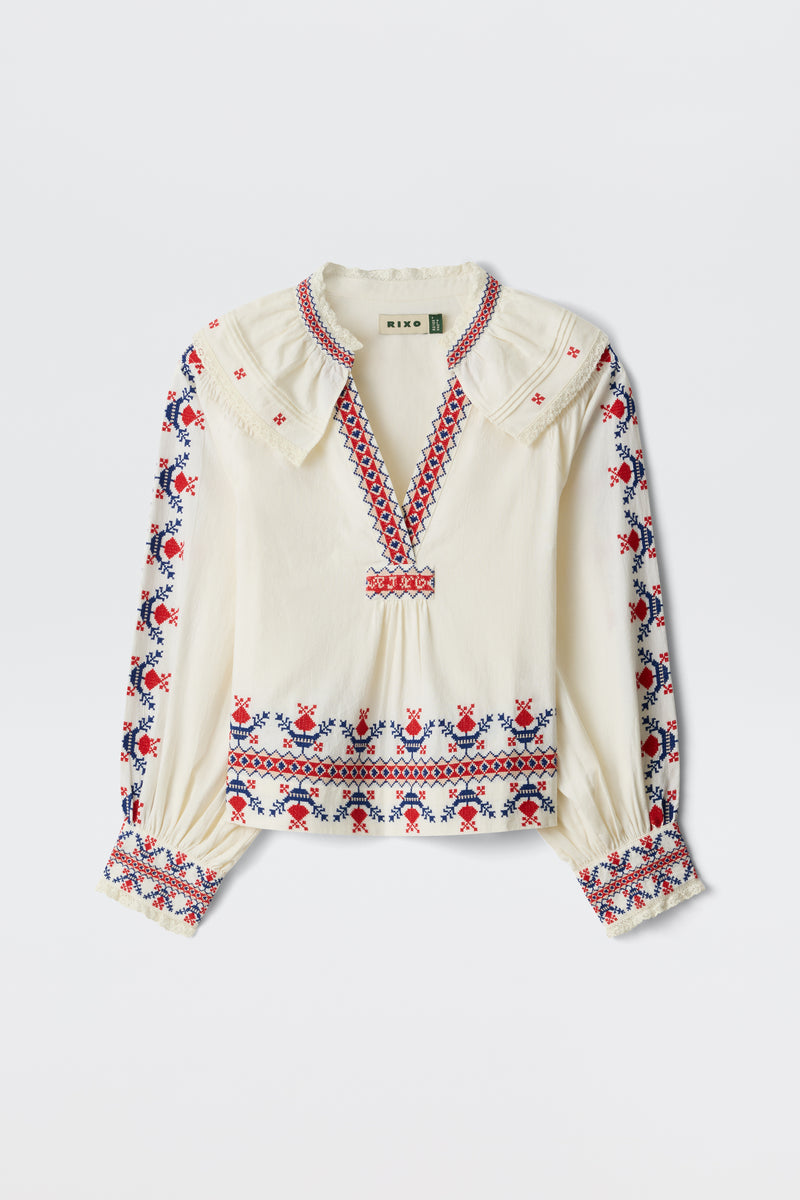 The Brisella - Gea Geo Embroidery Cream blouse features a cream color, long sleeves, and red and blue floral embroidery on the collar, cuffs, front, and sleeves. It has a V-neck with a slightly ruffled collar.