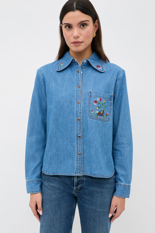 A woman faces forward wearing the Edaline Sadie Embroidery Light Vintage denim shirt, featuring colorful embroidery on the collar and chest pocket, paired with blue jeans against a plain light background.