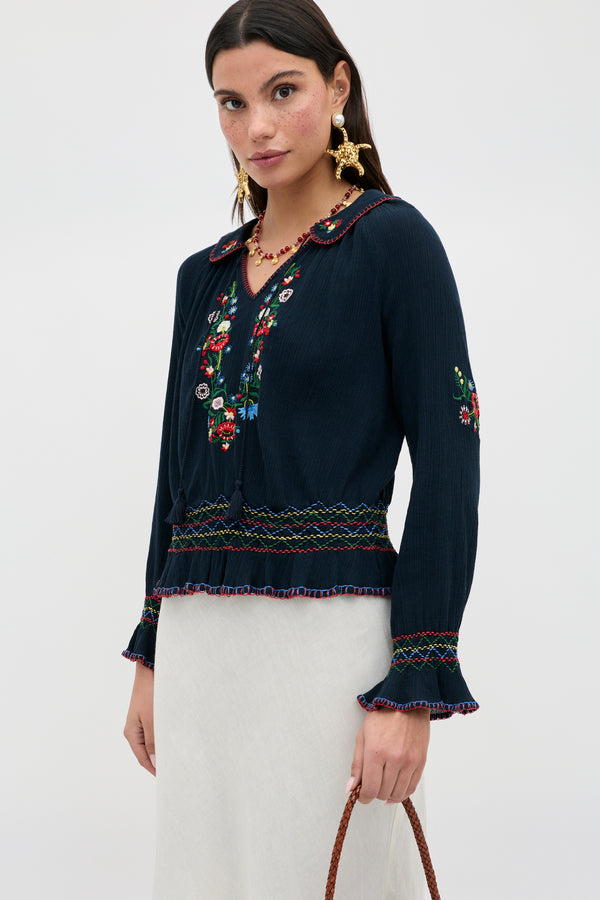 A woman wears the Sofira - Petunia Embroidery Navy blouse with floral designs, ruffled cuffs, and a scalloped collar, paired with a light skirt and holding a brown woven bag against a plain background.