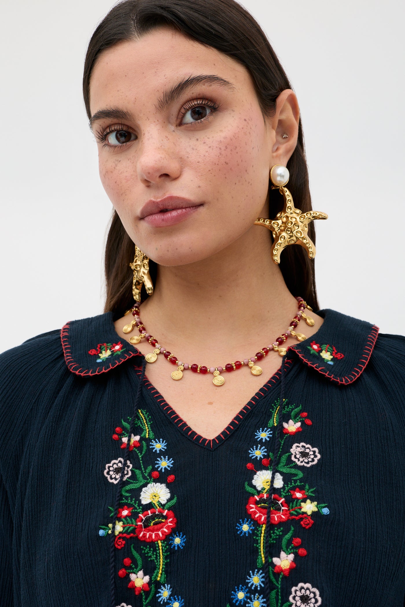 A woman with long brown hair wears the Sofira - Petunia Embroidery Navy top. She pairs it with gold starfish earrings, a beaded gold necklace, and gazes slightly past the camera against a plain background.