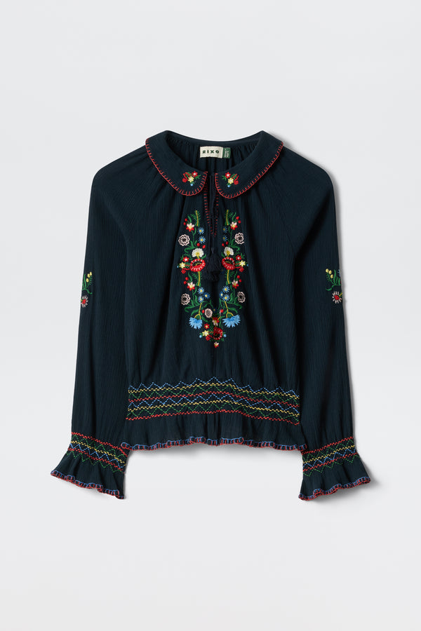 The Sofira - Petunia Embroidery Navy is a dark, long-sleeved blouse with colorful floral embroidery on the front, sleeves, and hem, featuring a Peter Pan collar, ruffled cuffs, and multicolored decorative stitching.