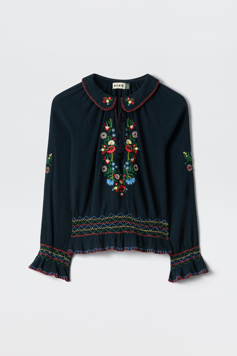 The Sofira - Petunia Embroidery Navy is a dark, long-sleeved blouse with colorful floral embroidery on the front, sleeves, and hem, featuring a Peter Pan collar, ruffled cuffs, and multicolored decorative stitching.