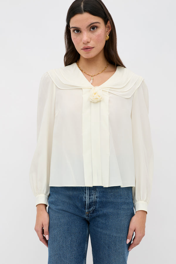 A woman wearing the Emelin - Ivory blouse, featuring long sleeves, a sailor-style collar, and a floral detail at the neckline, paired with blue jeans against a plain white background.